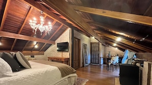 A cozy attic bedroom with a large bed, a chandelier, a TV, and a stone fireplace.