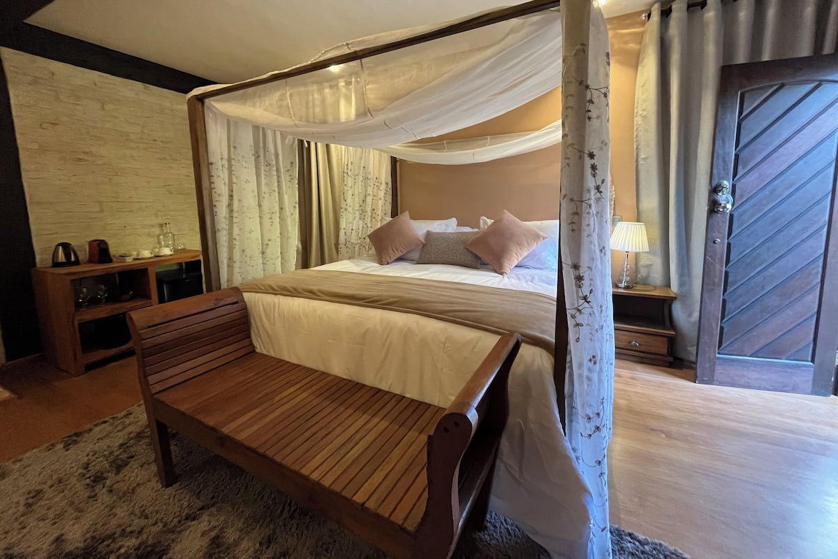 Una cama con dosel, un banco de madera, una mesita de noche con lámpara y una puerta con un diseño geométrico.