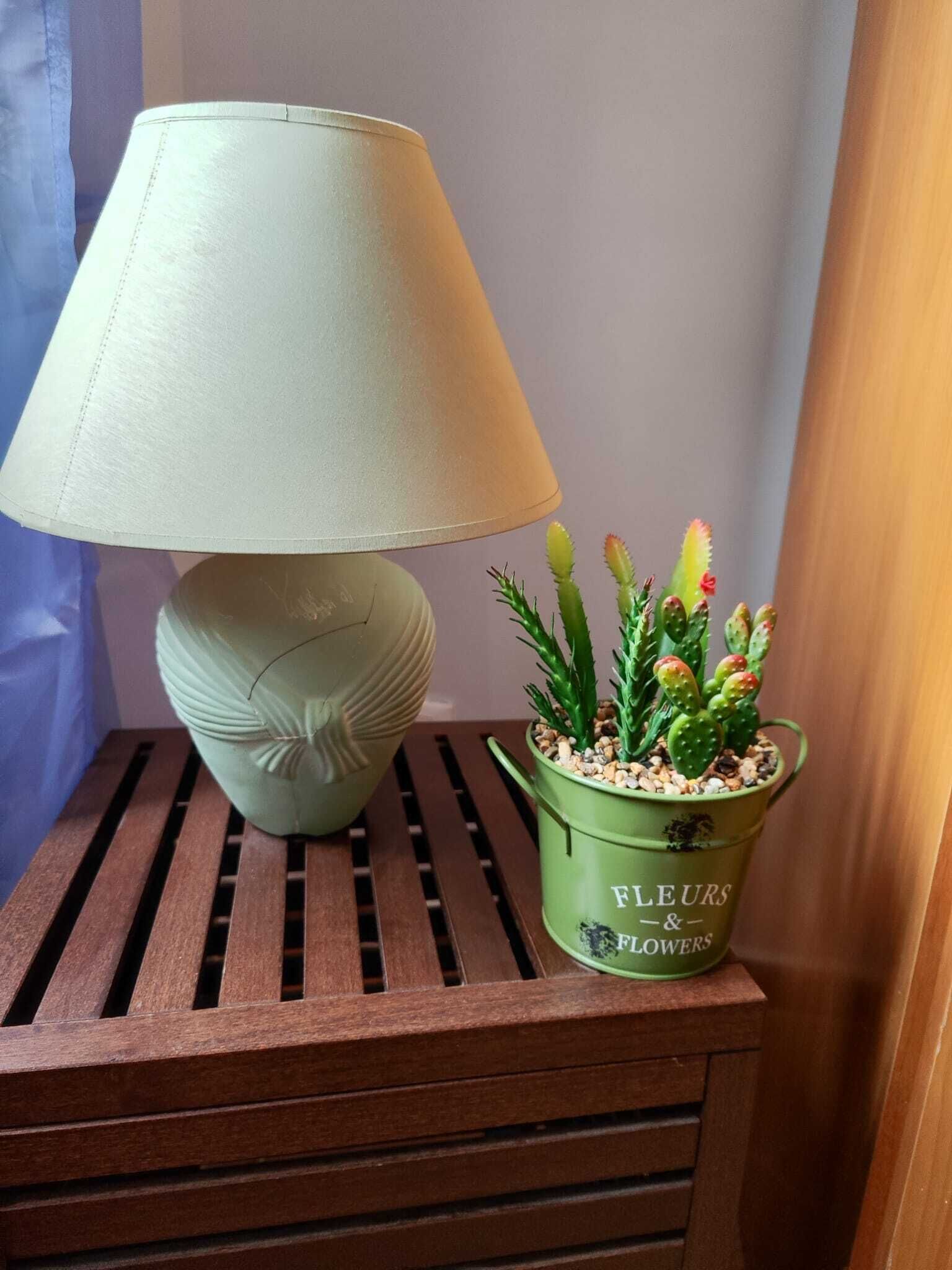 En lampa med en beige lampskärm och en grön kruka med texten ”FLEURS & FLOWERS” på ett träbord bredvid sängen.