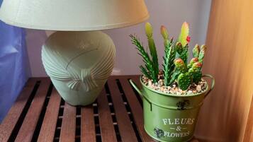 En lampa med en beige lampskärm och en grön kruka med texten ”FLEURS & FLOWERS” på ett träbord bredvid sängen.