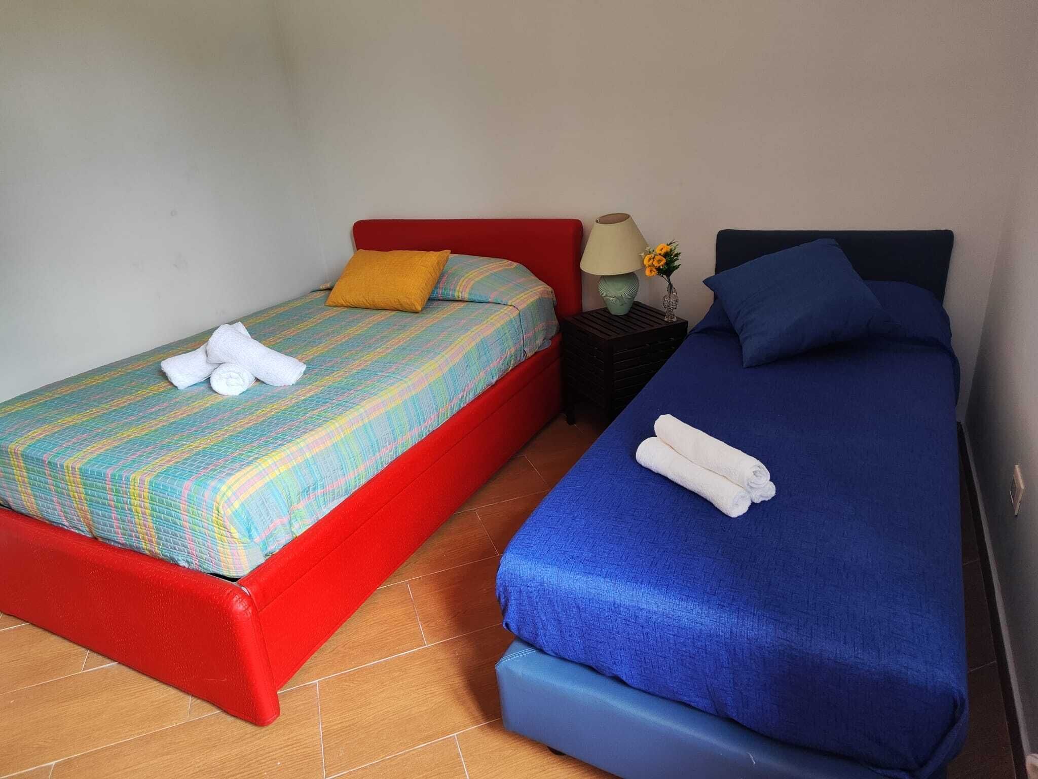 Two single beds with colorful bedding, a small table with a lamp and flowers, and a red and blue headboard.