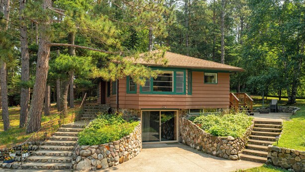 A two-story cabin with a stone staircase leading to the entrance, surrounded by a forested area.