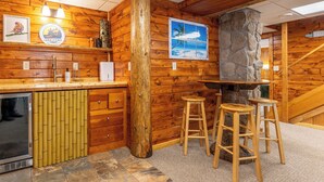 A wooden cabin interior with a bar area, stools, a stone fireplace, and a view of the outdoors.
