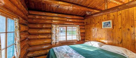 Una camera in una baita di legno con un letto, due finestre e un quadro appeso al muro.