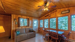 A cabin interior with a wooden ceiling, a ceiling fan, a sofa, a dining table with chairs, and large windows offering a view of trees. - Lakefront Cabin with Shared Dock (Orr)