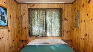 A wooden cabin room with a bed, a window with floral curtains, and a wall-mounted light switch. - Lakefront Cabin with Shared Dock (Orr)