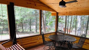 A wooden cabin interior with a swing chair, a round metal table with four chairs, and large windows offering a forest view. - The Most Private, Pet-Friendly Cabin (New Screened Porch in 2024) (Orr)