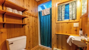 A wooden bathroom with a toilet, shelves with toilet paper, a window with curtains, a sink, and a shower curtain. - The Most Private, Pet-Friendly Cabin (New Screened Porch in 2024) (Orr)