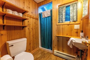 Une salle de bain en bois comprenant des toilettes, des étagères avec du papier toilette, une fenêtre avec des rideaux, un lavabo avec une serviette et un rideau de douche.