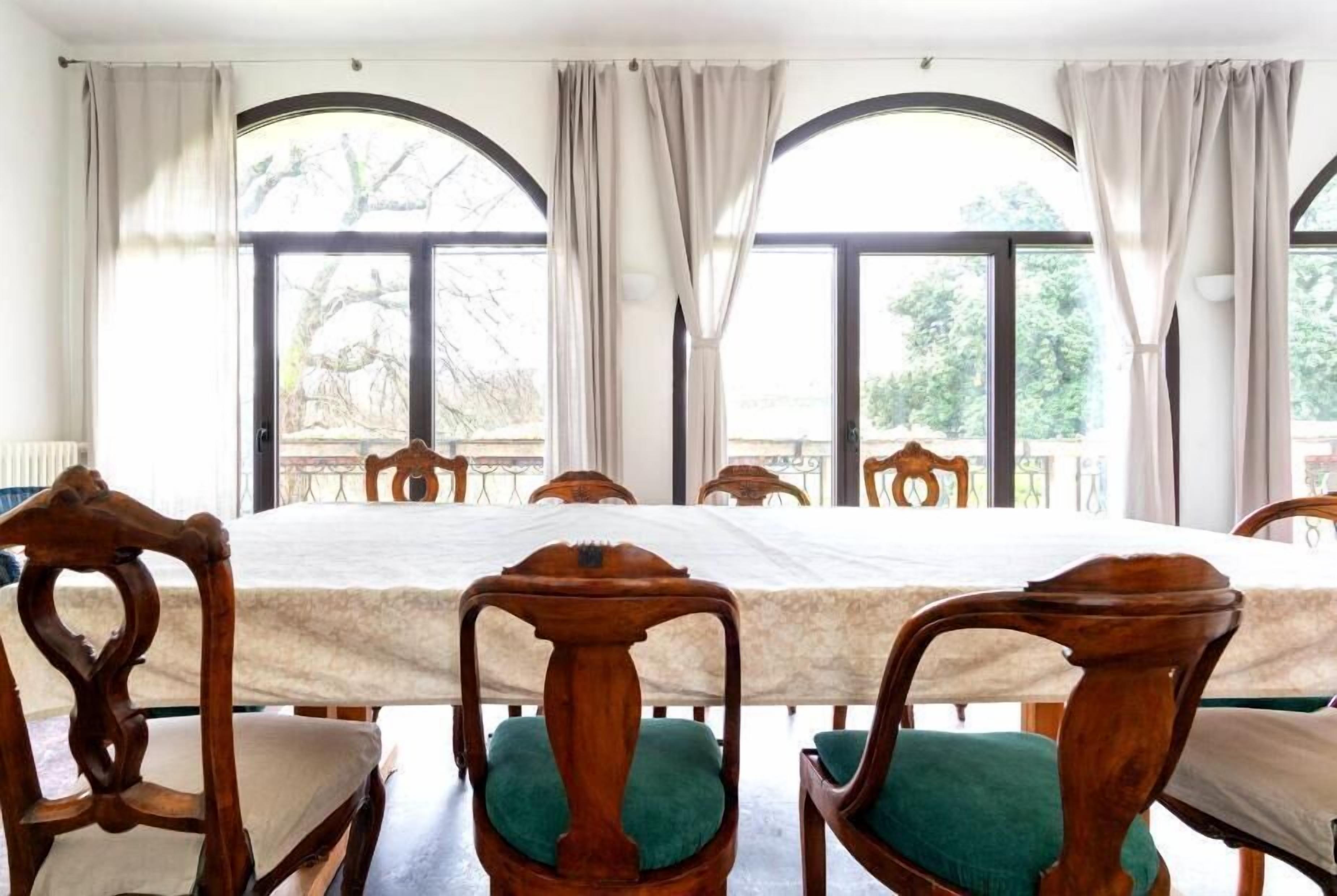Un espace repas avec une grande table et des chaises en bois, de grandes fenêtres avec des rideaux et une vue sur les arbres à l’extérieur.