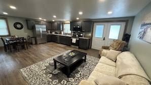 A modern kitchen with dark cabinets, a dining area with a wooden table and chairs, and a living room with a beige sofa and patterned rug.