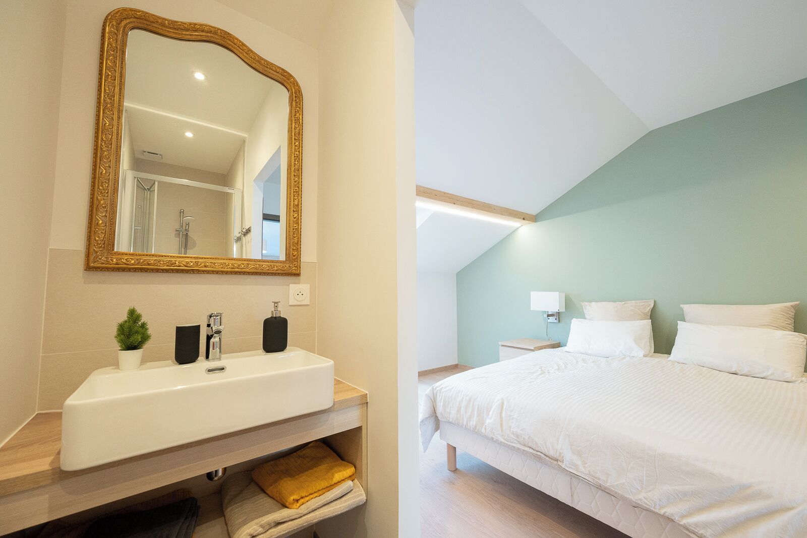 A modern bathroom with a large mirror, white sink, and a small potted plant. The bedroom features a large bed with white bedding and a light green wall.