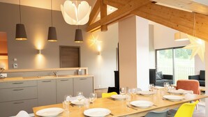 A modern dining area with a wooden table set for six, grey and yellow chairs, and a kitchen in the background with grey cabinets and pendant lighting.
