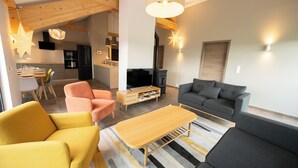 A modern living room with a sofa, armchairs, and a TV. There is a kitchen area with a dining table and chairs.