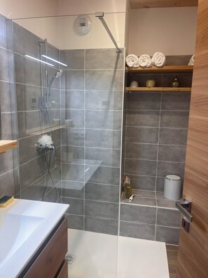 A modern bathroom with a glass-enclosed shower, grey tiles, a wooden shelf with towels, and a white sink.