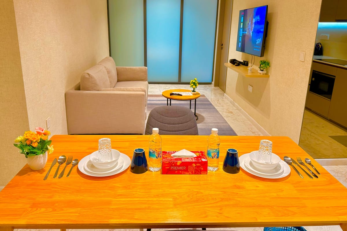 A dining area with a wooden table set for two, a sofa, a small round table with a plant, and a TV mounted on the wall.
