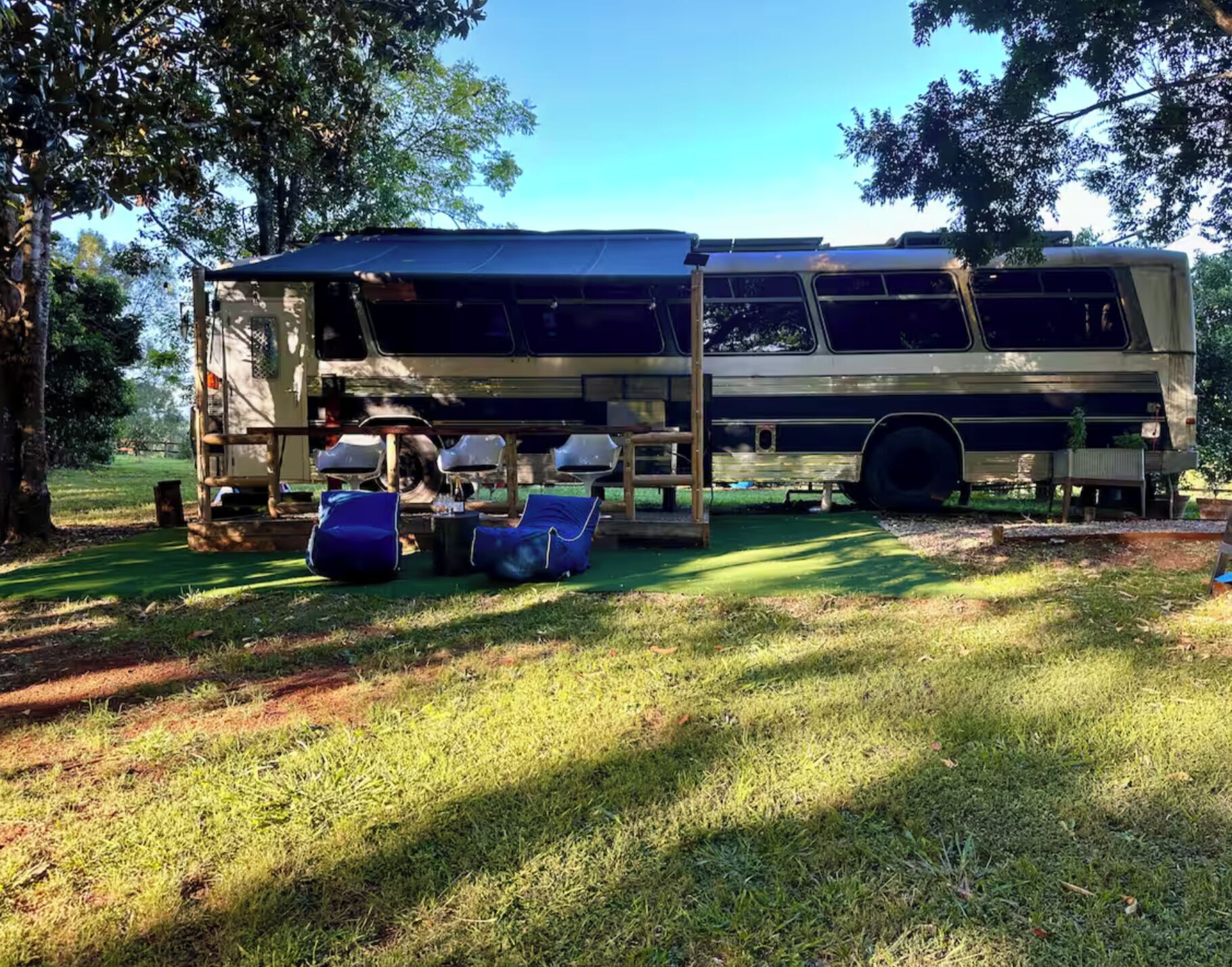 Ein Bus mit überdachtem Bereich und Sitzgelegenheiten, geparkt in einer grünen Umgebung mit Bäumen.