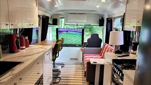 A compact living area with a kitchen, a TV displaying a sports game, and a window with a view of greenery. - The Bello BigRig - Luxury Riverfront Bus Conversion, close to town, pet friendly (Bellingen)
