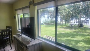 A room with a TV on a wooden stand, a window with a view of a grassy yard and trees, and a dining area with chairs and a table.