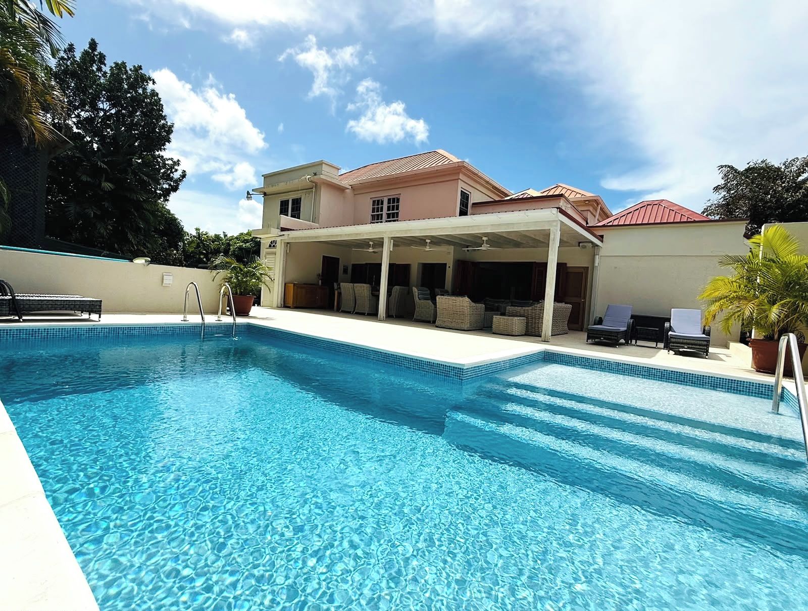 A swimming pool with a covered patio area, lounge chairs, and a house in the background.