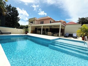 A swimming pool with a covered patio area, lounge chairs, and a house in the background. - Jack's Place - by Zenbreak (Mullins)
