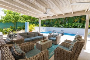 A covered outdoor seating area with wicker furniture, a pool, and lush greenery in the background. - Jack's Place - by Zenbreak (Mullins)
