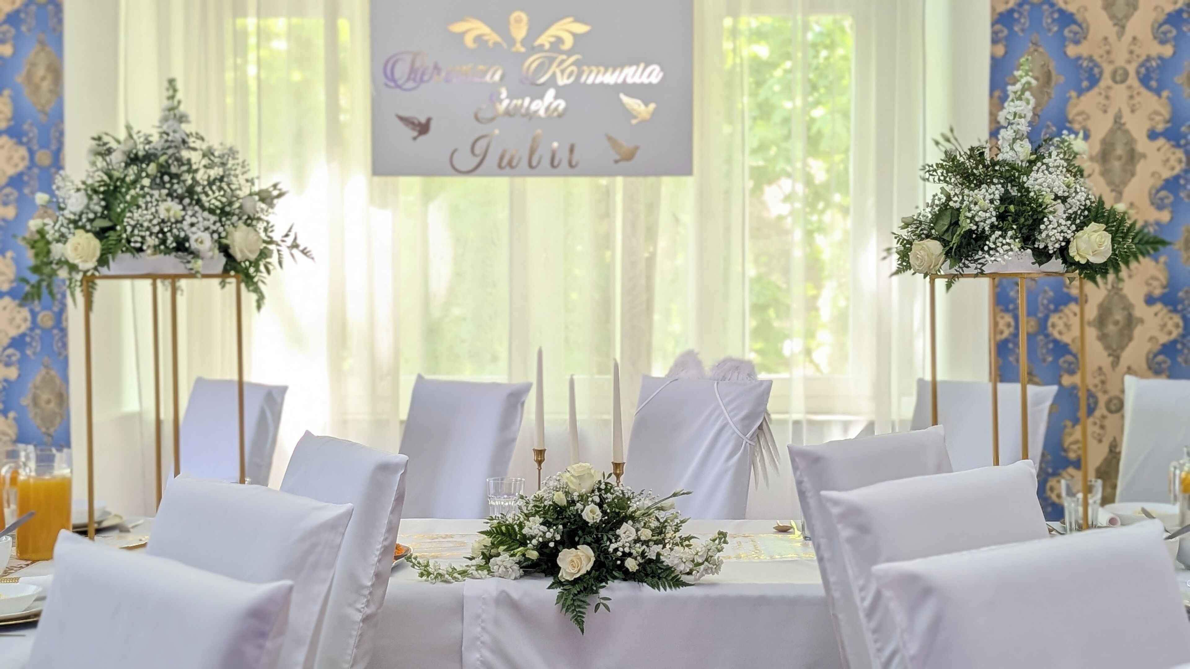 Et festbord til et bryllup med hvide stole, blomsterarrangementer og en banner med tekst.