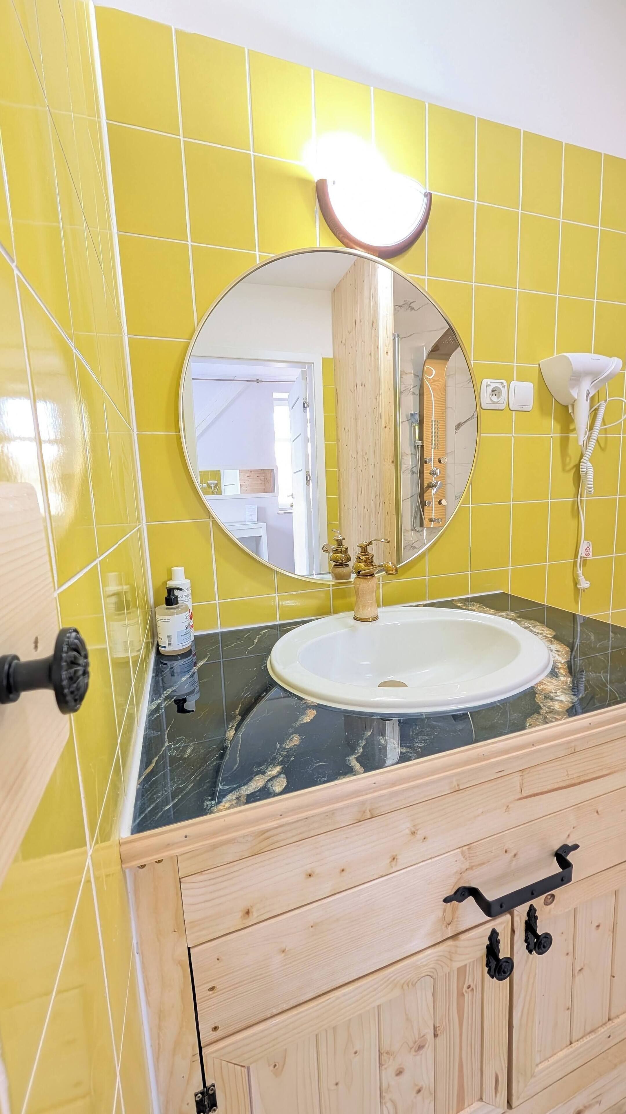 A bathroom with yellow tiles, a round mirror, a white sink, and wooden cabinets.