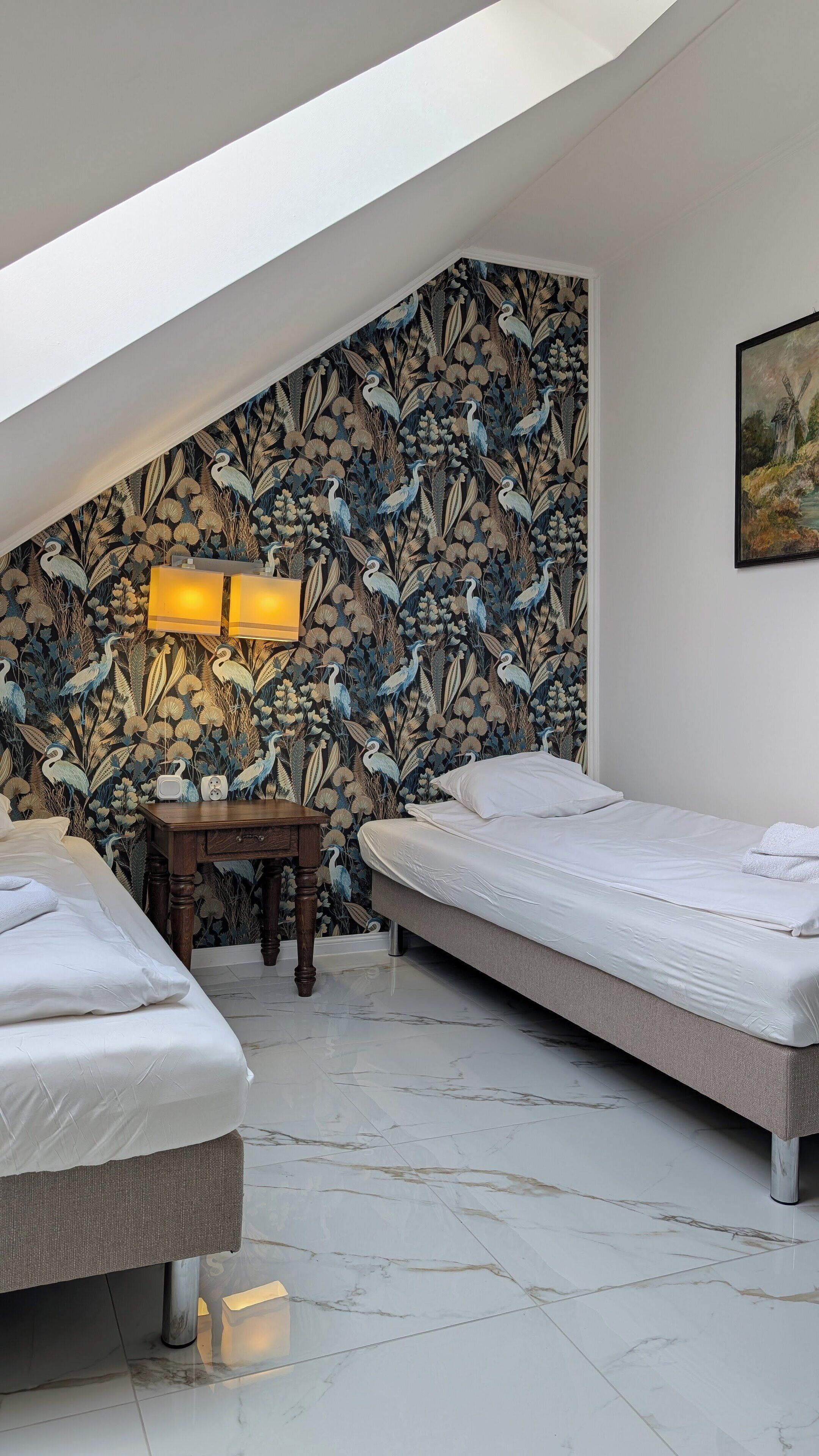A bedroom with two beds, a wooden nightstand, a wall-mounted lamp, and a painting.