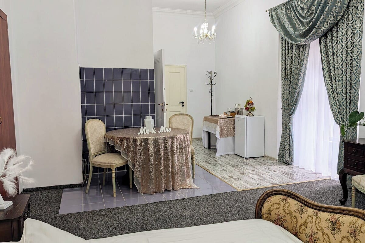 A hotel room with a bed, a desk with a chair, a small table with a chair, a chandelier, and curtains.