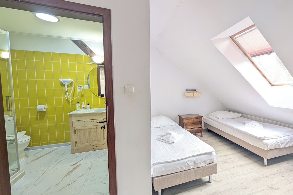 A bedroom with two beds, a small wooden dresser, a window with blinds, and a bathroom with a yellow wall and a mirror.