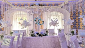 Een luxueuze balzaal met elegante witte en gouden decoratie, waaronder kroonluchters, bloemstukken en een centraal geplaatste bloemstelling in de vorm van een hart.