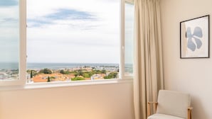 Habitación con un ventanal grande que ofrece vistas a un pueblo costero, una silla y una obra de arte abstracto enmarcada en la pared.