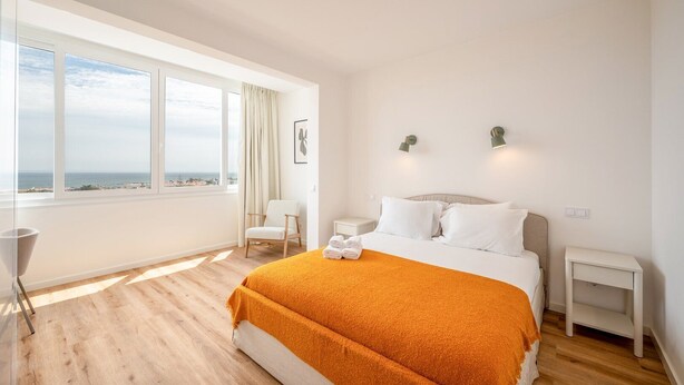Un dormitorio con un ventanal grande, una cama con manta naranja, una silla y una mesita de noche.