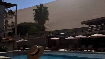 A pool area with lounge chairs and umbrellas, a palm tree, and a building with a desert landscape in the background.