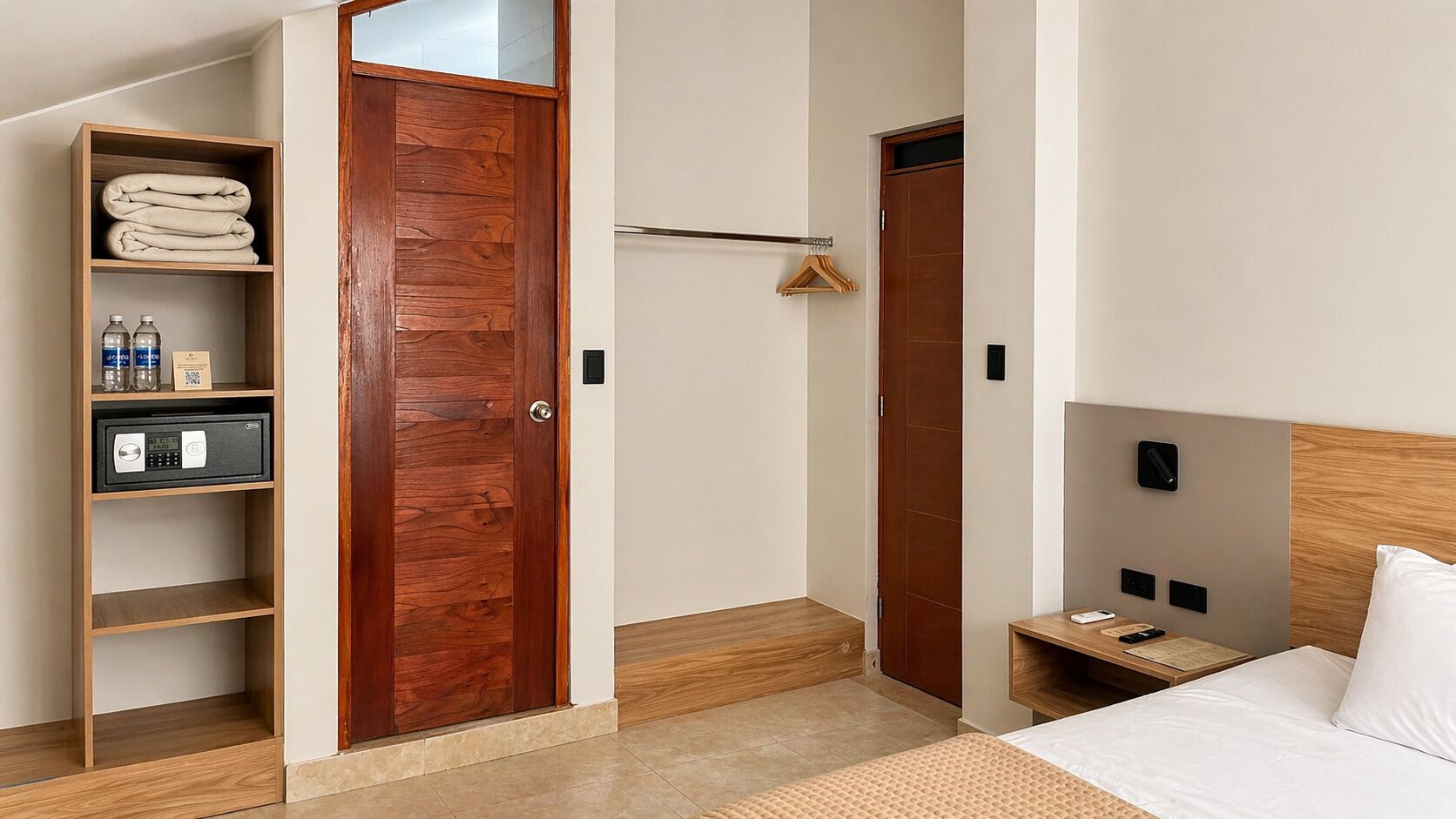 A hotel room with a bed, a wooden wardrobe, a small table with a remote, and a shoe rack.