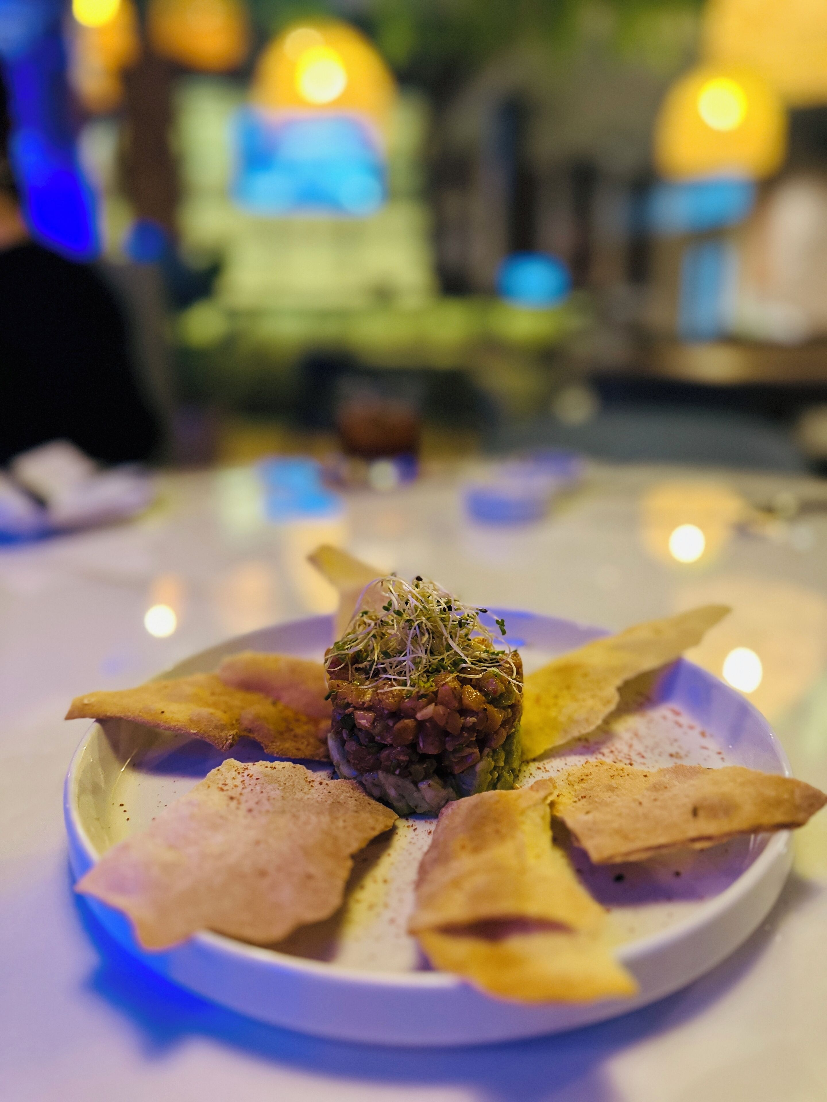 Un plato de comida con una porción de ingredientes en el centro, rodeado de panes planos, sobre una mesa con un fondo borroso de luces.