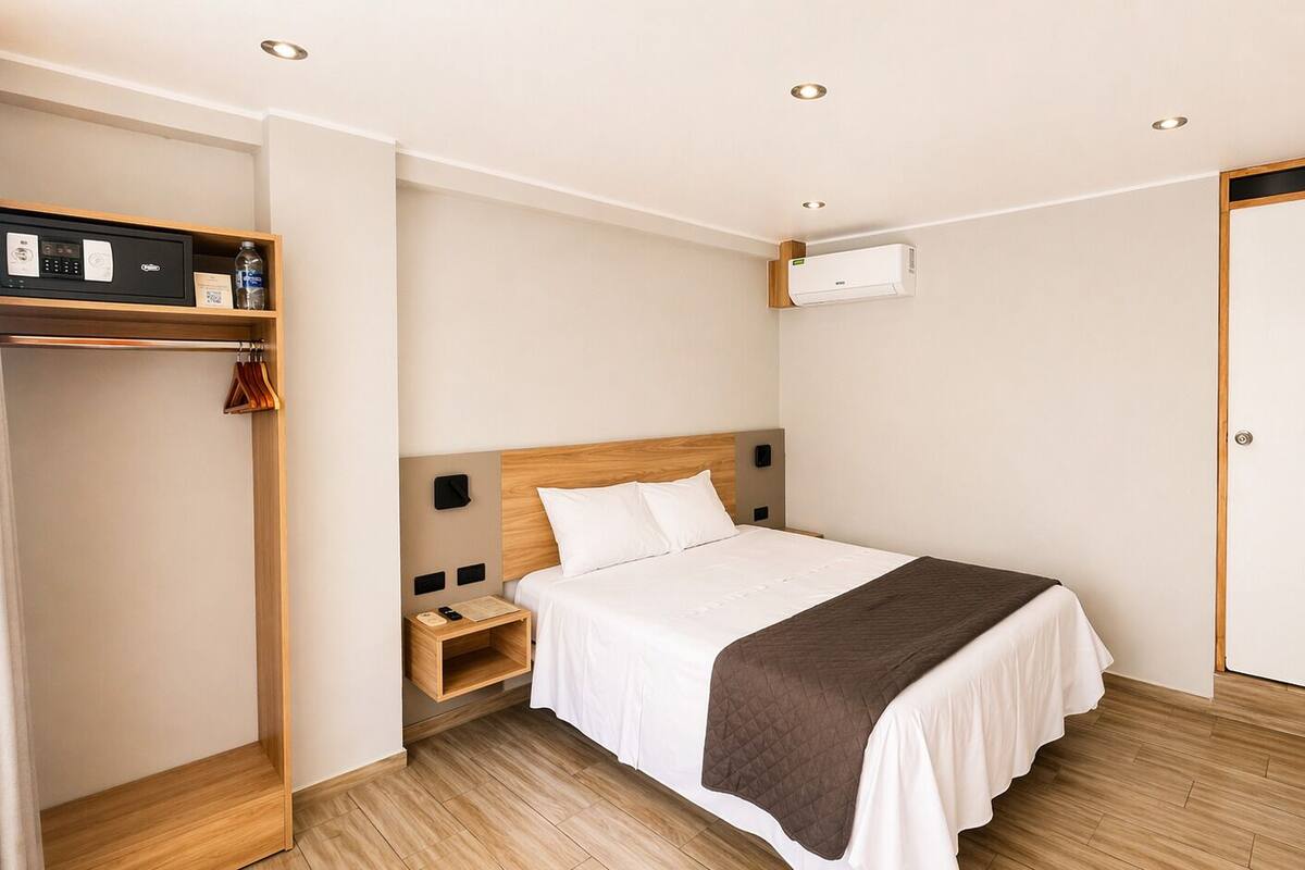 A hotel room with a bed, bedside table, nightstand, wardrobe, and an air conditioning unit.