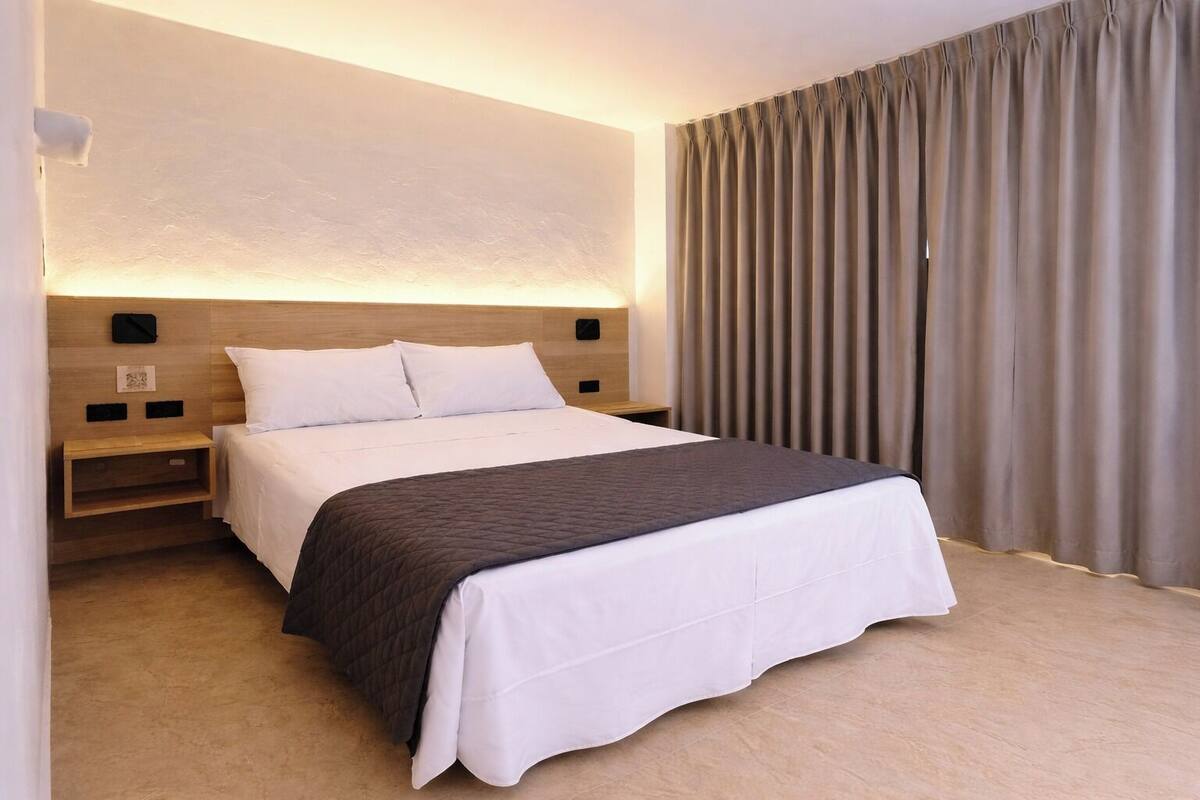 A hotel room with a large bed, a bedside table, a nightstand, a wall-mounted lamp, and a window with curtains.