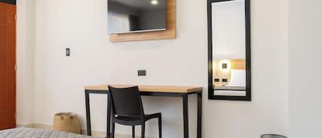 Un dormitorio con cama, escritorio, silla, televisor, espejo y cubo de basura.