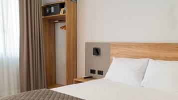 Habitación de hotel con una cama grande, cabecera de madera, mesita de noche, armario y ventana con cortinas.