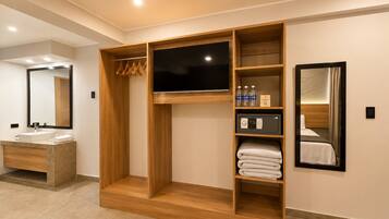 A modern hotel room with a wooden wardrobe, a flat-screen TV, a minibar, and a large mirror.