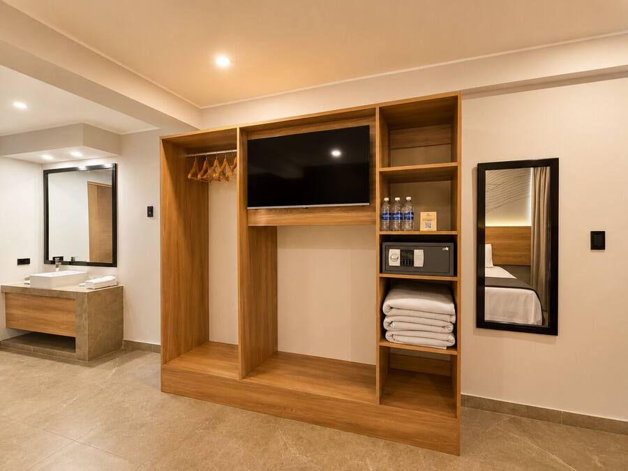 A modern hotel room with a wooden wardrobe, a flat-screen TV, a minibar, and a large mirror.