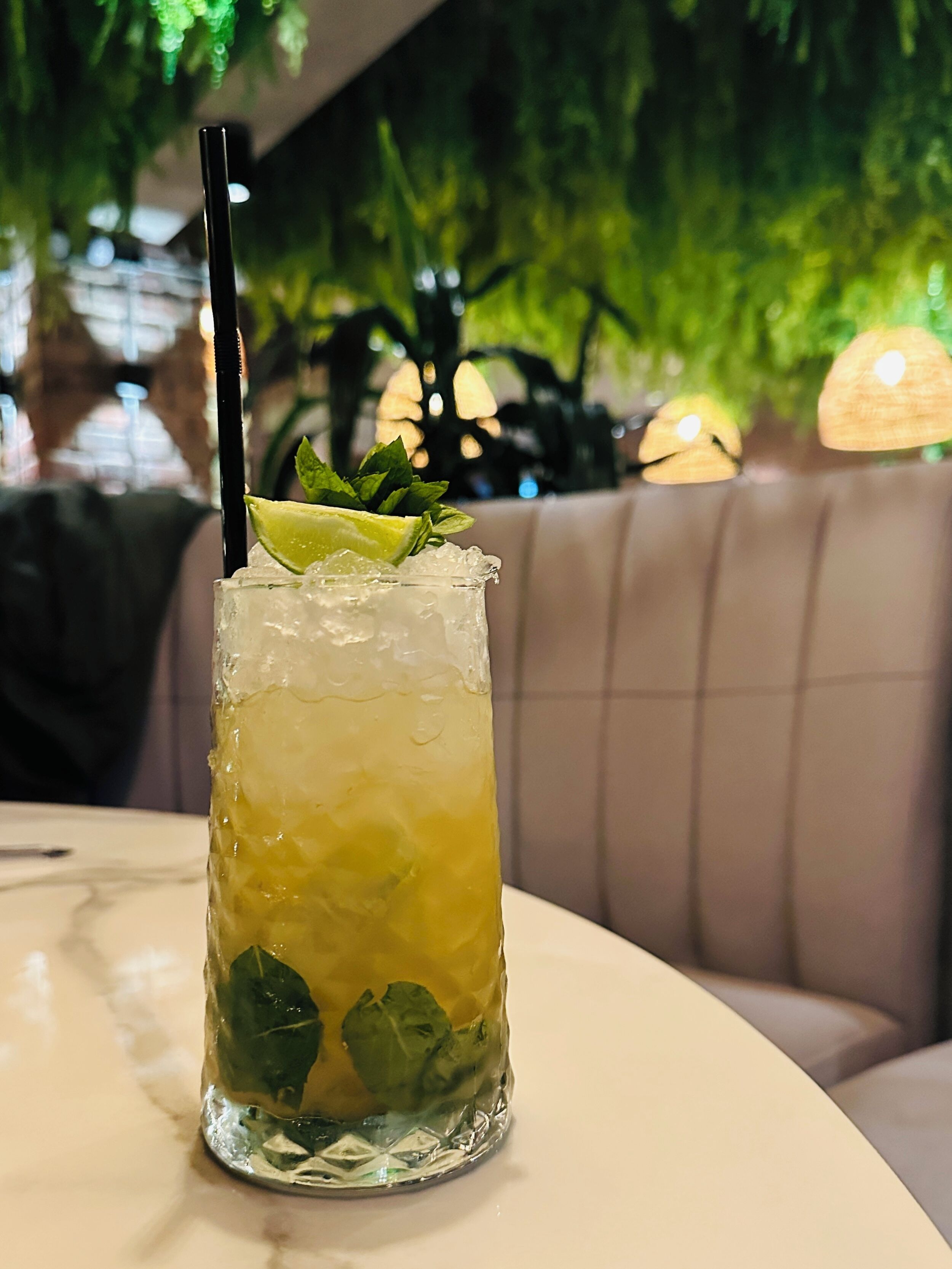 Un vaso alto con un gajo de lima y hojas de menta, lleno de hielo y un refresco amarillo, sobre una mesa en un restaurante con un fondo verde y frondoso.