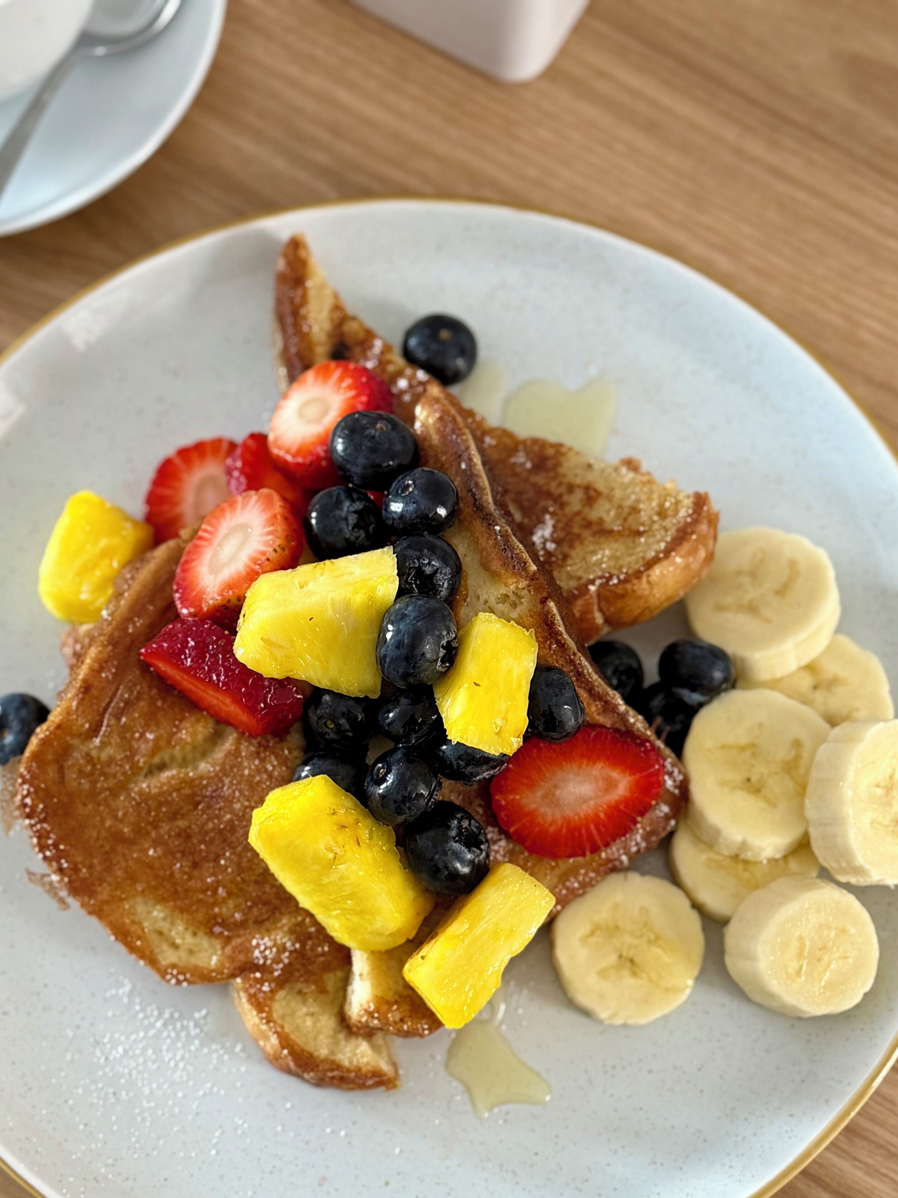 A plate of French toast topped with sliced bananas, strawberries, blueberries, and pineapple chunks.