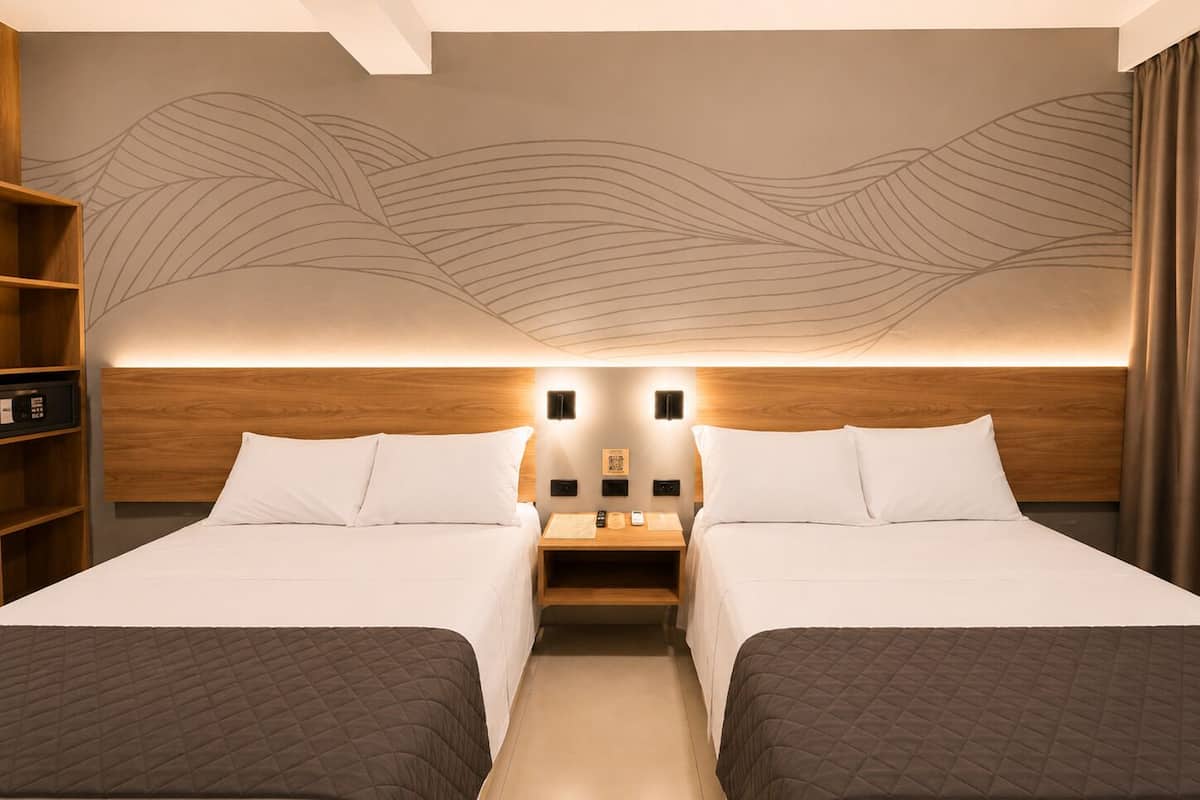 A hotel room with two beds, a wooden headboard, a wall mural, bedside tables, and a bookshelf.