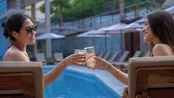 Dos personas brindando con bebidas junto a una piscina, con sillones de descanso y sombrillas al fondo.