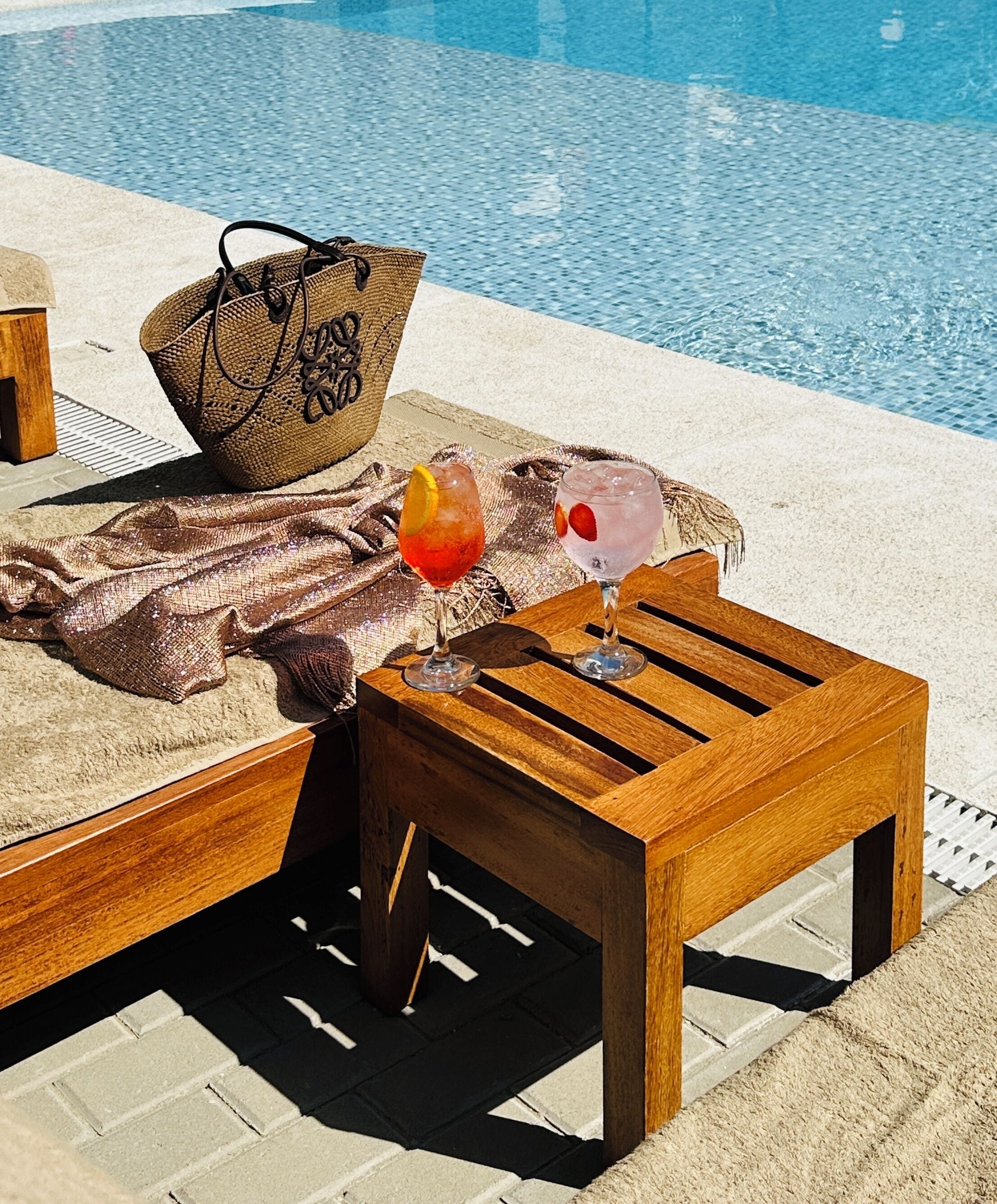 A poolside area with a wooden table, two drinks, a beach bag, and a towel.