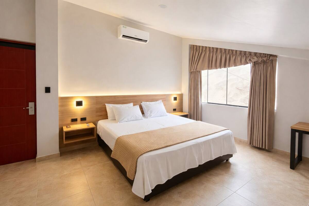 A hotel room with a bed, bedside tables, an air conditioning unit, a window with curtains, and a red door.