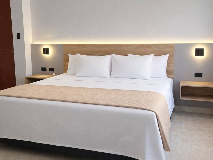 Een net opgemaakt bed met witte lakens en een beige plaid, met aan weerszijden nachtkastjes met ingebouwde verlichting, in een moderne hotelkamer.